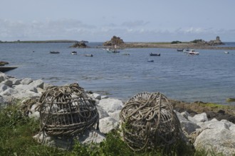 Rolled fishing baskets on a rocky coastline with boats and calm waters, St Agnes, Isles of Scilly,