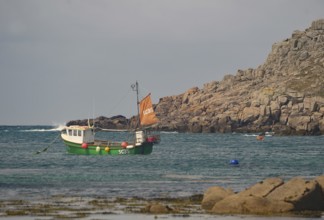 Small boat with orange sail in calm coastal waters near rocky cliff, Tresco, Isles of Scilly,