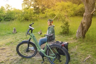 Cyclist with helmet sitting relaxed next to a bicycle in a green, summery environment, e-bike
