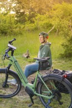 Person with helmet sitting in the sunset next to a bicycle in a meadow, surrounded by nature,