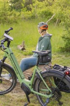 A person wearing a bicycle helmet sits on a tree stump next to a bicycle in the forest, e-bike