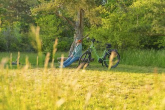 Cyclist sitting relaxed next to a bicycle under a tree on a green meadow, e-bike excursion,