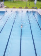 A person in a turquoise-coloured swimming pool swims laps surrounded by empty lanes on a warm