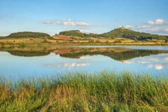 Wachsenburg Castle is reflected in the still water of a lake under a blue sky with cumulus clouds,