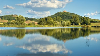 Wachsenburg Castle is reflected in the still water of a lake under a blue sky with cumulus clouds,