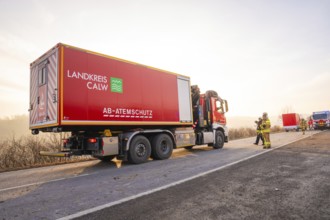 A large red fire truck is parked on a landscape road in the morning light, firefighters next to it,