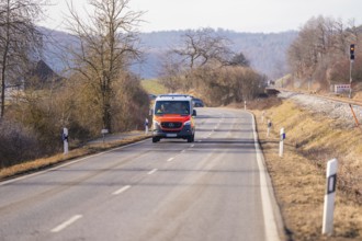 An ambulance is driving on a rural road, trees and railroad tracks in the background, fire