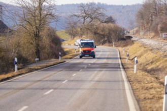 An ambulance is driving on a rural road. Sparse vegetation and trees surround the road, fire