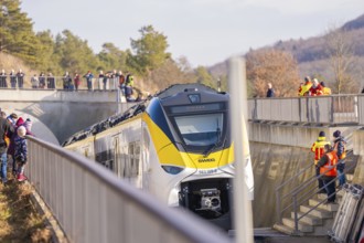 A yellow and white train travels under a bridge with spectators. Staff wearing safety vests