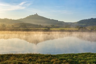Wachsenburg Castle is reflected in the still water of a lake in the morning light, fog rises,