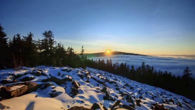 The sun sets behind a snow-covered mountain, surrounded by trees, thick fog in the valley,