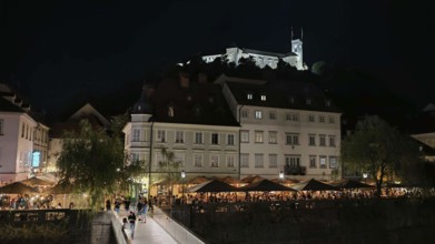 Castle illuminated at night surrounded by illuminated restaurants and visitors in an urban setting,