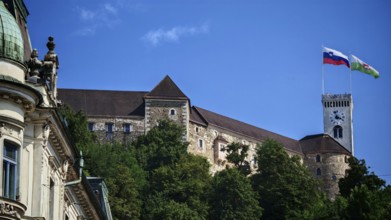 Mighty castle with two flags on a tower nestled in green trees against a clear sky, Ljubljana,