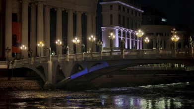 Illuminated bridge over a river at night with urban architecture in the background, Skopje, North
