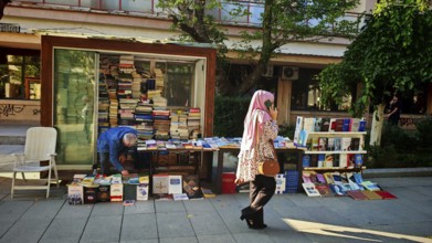 Book stand on a street surrounded by people and green trees, idyllic street scene with woman