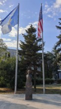 Bust statue of Madeleine Albright outdoors with two USA and Kosovo flags surrounded by tall trees,