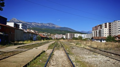 Overgrown train tracks run through a town at the foot of mountains on a sunny day, Kosovo
