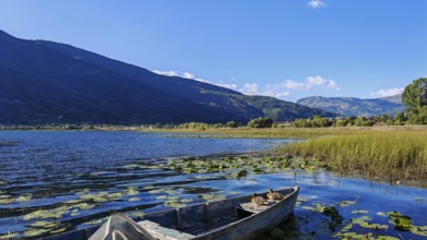 A boat on a lake with surrounding reeds and mountains in the background under a blue sky, Lake