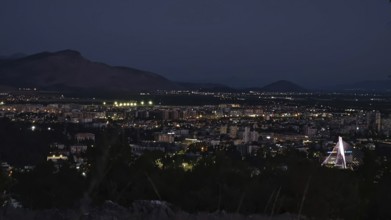 Nighttime city view with illuminated buildings, mountains in the background and dark sky,