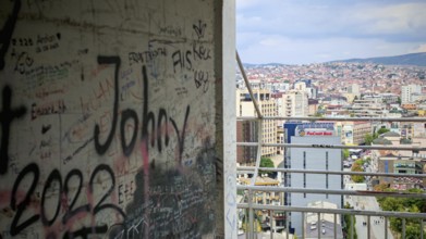 A graffiti-covered building offers a view of an urban landscape under a cloudy sky, Pristina,