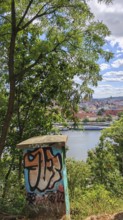 View of a city and a river through trees, in the foreground a power box covered with graffiti,