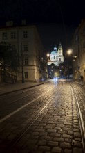 Illuminated cobblestone street with tram tracks at night with a view of a cathedral, Prague, Czech