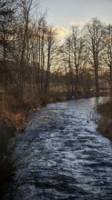 A quiet river flows through a winter landscape lined with bare trees, Franconian Forest nature park