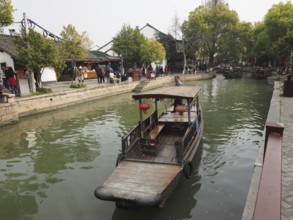 A wooden boat floats on a canal next to traditional buildings and market visitors, Zhujiajiao