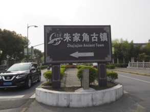 A road sign points to the ancient city of Zhujiajiao surrounded by trees and a road with cars,