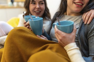 Two young women friends enjoying hot drinks and conversation while relaxing on a sofa at home,