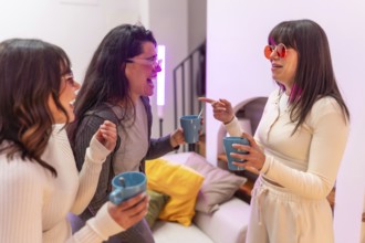 Three young women laughing and chatting over hot drinks in a cozy apartment, enjoying a relaxed