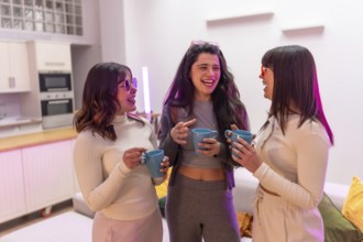 Three diverse young women laughing and talking while holding coffee mugs, gathering in a modern