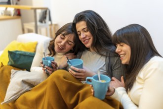 Three smiling young women enjoying a cozy moment together on a sofa at home, sharing drinks and