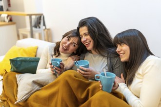 Three young women on a sofa wrapped in a blanket, sharing hot drinks, laughing and chatting in a