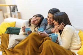 Three young women on a sofa sharing a cozy blanket, laughing and sipping warm drinks while enjoying