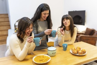 Three young women smiling and laughing, holding cups and spoons, gathering around a wooden kitchen