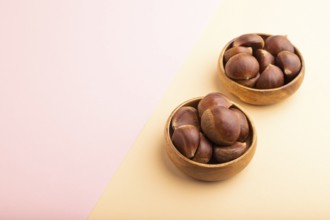 Wooden bowl with raw edible ?hestnuts on pink and orange paper pastel background, side view, copy