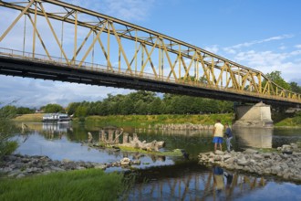 Yellow metal bridge across river with two anglers on the shore and blue sky, bridge over Morava