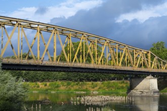 Yellow metal bridge over a river with green vegetation on the banks and cloudy sky, bridge over the