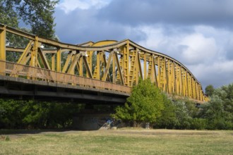 Yellow metal bridge over a meadow with trees, a river and cloudy sky in the background, bridge over