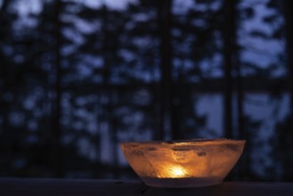 Ice bowl illuminated by burning tea light stands on balcony balustrade, forest at the onset of