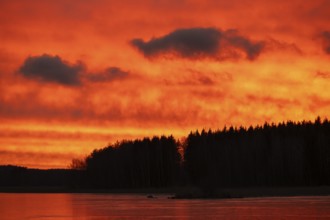 A dramatic sunset over a still, frozen lake and forest with orange-red hues in a cloudy sky,