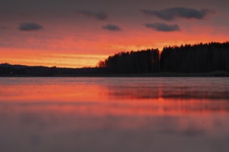 A calm sunset sky with orange-red-yellow hues over a quiet lake and dark forest, Finland
