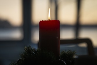 A single red candle in an advent wreath burns in front of a window at dusk and creates a cozy