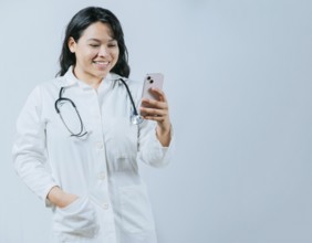 Female healthcare worker holding cellphone. Smiling asian doctor using smartphone isolated, Young