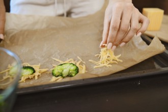 A person is placing shredded cheese on a baking sheet in a kitchen. The broccoli is ready to be