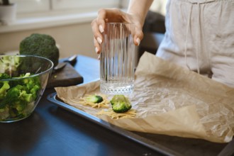 Woman is preparing smashed broccoli with cheese in a kitchen. She is using a glass to smash the
