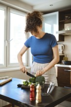 Woman chops broccoli in a kitchen. The cutting board is set on a table beside a bowl. Seasoning