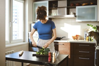 Woman is chopping broccoli on a black cutting board in a bright kitchen. The window shows daylight