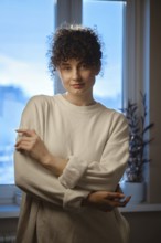 A young woman stands near a window in a dim room. She is wearing a light sweater and looks at the